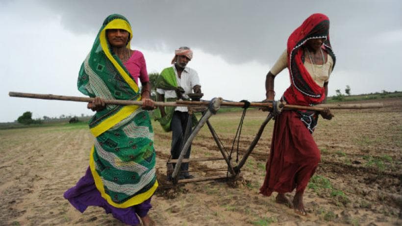 Two rural women pull a traditional plough on farmland as a man guides, highlighting women’s labour in Indian agriculture.