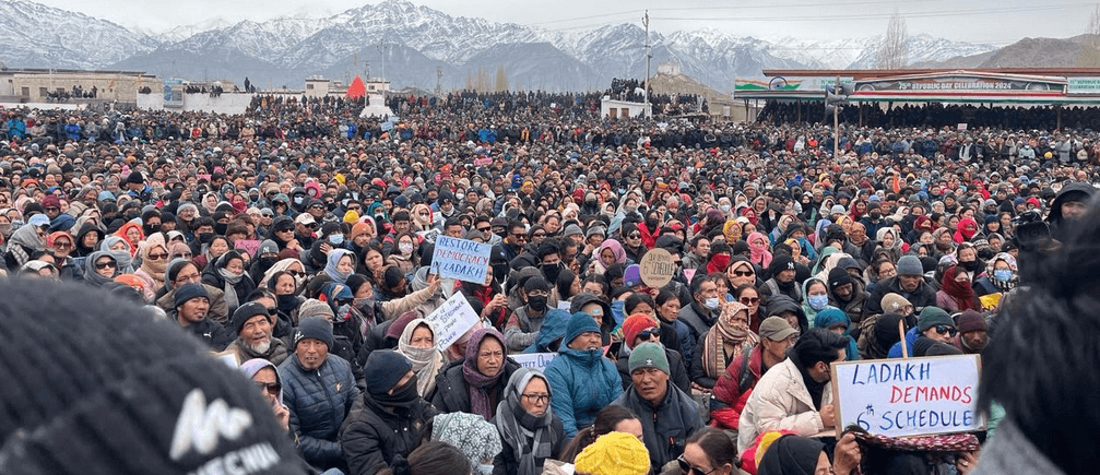 Large crowd in Ladakh protesting with banners demanding 6th Schedule rights, snowy mountains visible in the background.