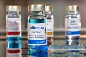 A vial labeled “Influenza vaccine” with a syringe in front and blurred vaccine bottles in the background