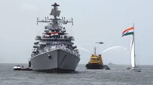 Indian Navy warship sails with support vessels during a ceremony, water jets and flag in view.